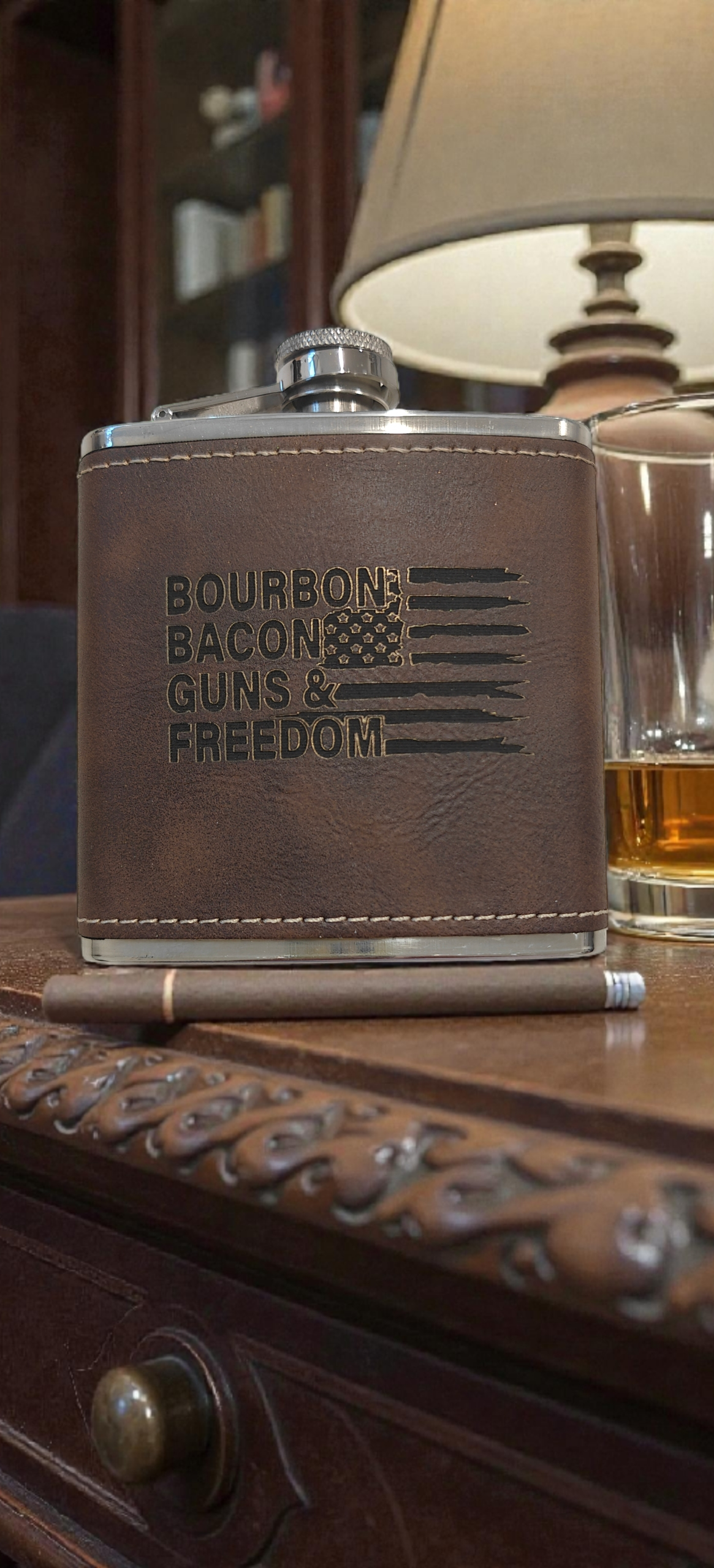 Flask with 'Bourbon Bacon Guns & Freedom' engraving on a leather surface.